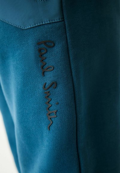 Modré teplákové nohavice majú hladký povrch a po strane je umiestnené čierne embosované logo "Paul Smith", čo posilňuje moderný dizajn.