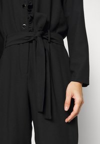 Combinaison noire à manches longues avec ceinture en tissu nouée à la taille et boutons décoratifs, montrant une partie de la main détendue d'une personne sur le côté.