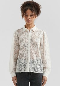 White lace blouse with button-up front, collared neck, and puff sleeves. Delicate floral pattern and transparent texture. Worn over dark jeans.