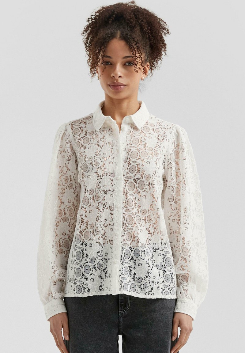 White lace blouse with button-up front, collared neck, and puff sleeves. Delicate floral pattern and transparent texture. Worn over dark jeans.