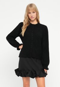 Svart, stickad cardigan med diamantmönster, rund halsringning och knappstängning. Har puffärmar och en avslappnad passform.