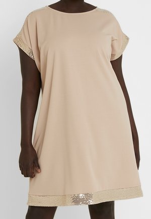 Vrouw draagt een losse beige jurk met korte mouwen, versierd met glinsterende paillettenbies aan mouwen en zoom.