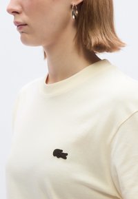 Μπεζ βαμβακερό μπλουζάκι με στρογγυλή λαιμόκοψη και κοντά μανίκια, με μικρό σκούρο καφέ κεντητό λογότυπο στην αριστερή πλευρά του στήθους.