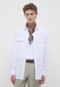 Camicia bianca con bottoni e due tasche sul petto, indossata sopra un top crema, con una sciarpa marrone a motivi e pantaloni verdi.