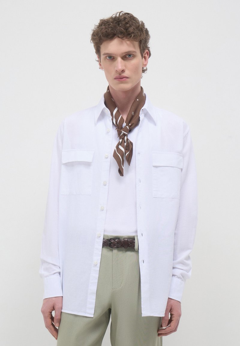 Camicia bianca con bottoni e due tasche sul petto, indossata sopra un top crema, con una sciarpa marrone a motivi e pantaloni verdi.