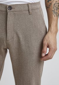 Beige kankaita trousers kanssa fain muotoilu, jossa on nappilukko ja sivutaskut, näytetään kädessä, joka lepäilee kankaan päällä.