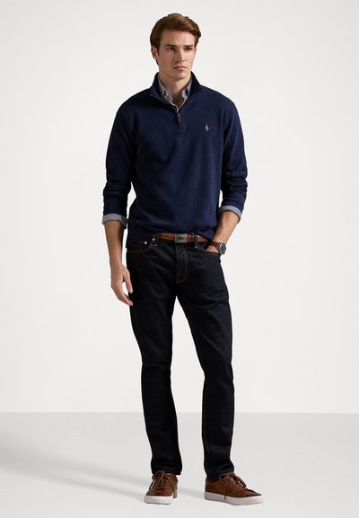 Young man wearing navy quarter-zip sweater, striped shirt, dark jeans, brown belt, watch, and brown sneakers, standing against plain background.