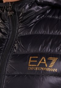Černá péřová bunda s celozipovým uzavřením a zlatým logem EA7 Emporio Armani na přední straně. Hladká, lesklá tkanina s prošívanou texturou.