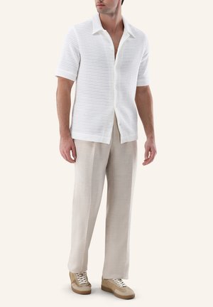 Mann trägt weißes strukturiertes Kurzarmhemd, hellbeige weite Hose und beige Sneakers, steht vor neutralem Hintergrund.