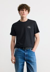 Νεαρός άνδρας με σγουρά μαλλιά φορώντας μαύρο μπλουζάκι με το λογότυπο Calvin Klein και μπλε τζιν με μαύρη ζώνη, στέκεται μπροστά σε απλό φόντο.