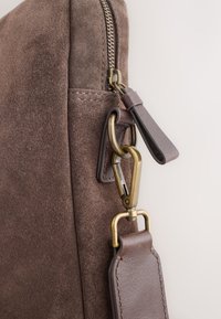 Brown suede bag side with brass zipper, leather zipper pull, and brass clasp attaching a leather strap.