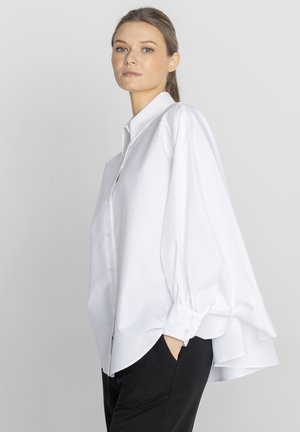 Kobieta ubrana w za dużą białą koszulę z pufiastymi rękawami i czarne spodnie, stojąca z jedną ręką w kieszeni na szarym tle.