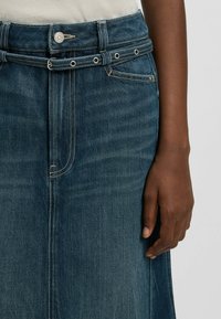 Tummansininen farkkuhame suoralla leikkauksella, jossa on nappi- ja lenkkikiinnitys, metallikoristeet ja näkyvät ompeleet.