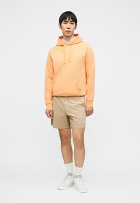 Άνδρας όρθιος, φοράει ροδακινί φούτερ με κουκούλα, μπεζ σορτς, λευκές κάλτσες και λευκά αθλητικά παπούτσια, σε απλό γκρι φόντο.