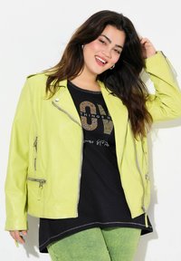 Veste de biker en cuir vert lime avec des accessoires en argent, poches zippées, superposée sur une chemise noire avec des détails en strass et un pantalon vert.