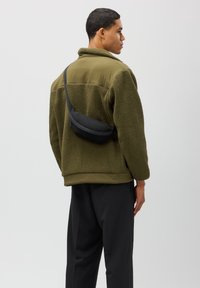 Fleece bunda v olivově zelené barvě s texturovaným povrchem, doplněná malou černou krosbody taškou. Černé kalhoty dotvářejí outfit.