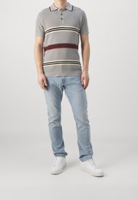 Camisa de polo de punto gris con cuello, que presenta tres botones y rayas horizontales en rojo, crema y negro. Combinada con jeans de color azul claro y zapatillas blancas.