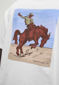 Camiseta blanca con una gráfica de un vaquero montando un caballo marrón contra un cielo azul claro y un fondo beige. Incluye el texto "originals studio '90".