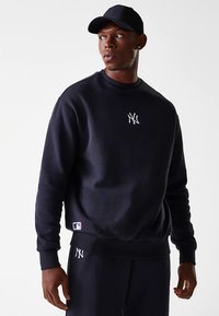 Mladý muž v čiernom mikinovom svetri New York Yankees, s rovnakými nohavicami a čiernou baseballovou čiapkou, sa pozerá doľava.