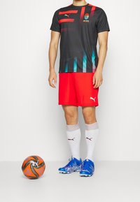 Černý a červený sportovní top s barevnými akcenty, červené šortky, modré kopačky, bílé ponožky se pruhy a oranžový fotbalový míč.