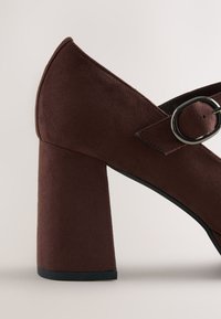 Brun mockaskor med hög klack och bred klack i blockstil, med justerbar rem och rund spänne. Slät textur och spetsig tådesign.