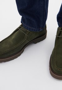Pantofi din suede verde cu un guler capitonat, având un design cusut și o talpă din cauciuc. Asociați cu o pereche de pantaloni negri din denim.