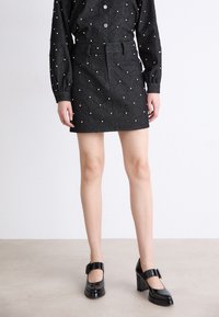Svart denim mini skjørt med teksturert diamantmønster og perlepynt, kombinert med svarte lakk Mary Jane hæler med en kraftig hæl.