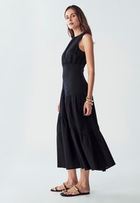 Musta, hihaton midi-mekko, jossa on rypytetty yläosa, kerroksellinen hame ja sileä pinta. Yhdistetty litteisiin sandaaleihin, joissa on kultaiset yksityiskohdat.