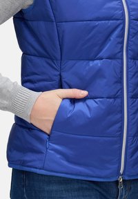 Blau, gefütterte Weste mit Reißverschluss, gestepptem Design und Seitentaschen. Graue Rippbündchen sind sichtbar. Leichtes, wasserabweisendes Material.