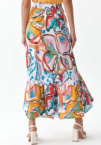 Žena v barevných květovaných sukni s volánovým lemem a bílých vysokých podpatcích, zobrazená ze zad.