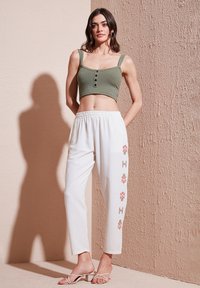 Oliwkowy prążkowany crop top z guzikami, zestawiony z białymi spodniami z kolorową haftowaną florystyką po bokach. Białe sandały na ramiączkach.