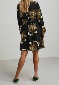 Abito nero floreale con maniche lunghe e vita elastica. Presenta una stampa botanica in crema e oro, con gonna a campana e orlo arrotondato.