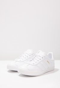 White leather sneakers with a smooth texture, featuring three horizontal stripes, round toe, and gold text accent. Classic rubber sole.