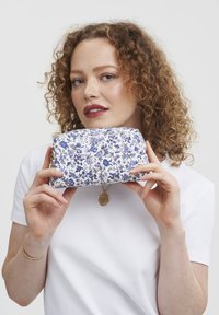 Pouch sa cvijetnim uzorkom u bijeloj i plavoj tkanini s zlatnim dijelovima. Ovalni oblik i teksturirana površina, držana u rukama protiv jednolične pozadine.