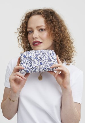 Floral-patterned zip pouch in white and blue fabric with gold hardware. Rounded shape and textured finish, held in hands against a plain background.