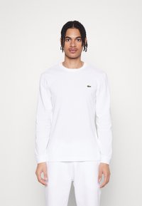 T-shirt blanc à manches longues en coton, avec un petit logo vert Lacoste sur la poitrine. Design simple à col rond. Texture douce.