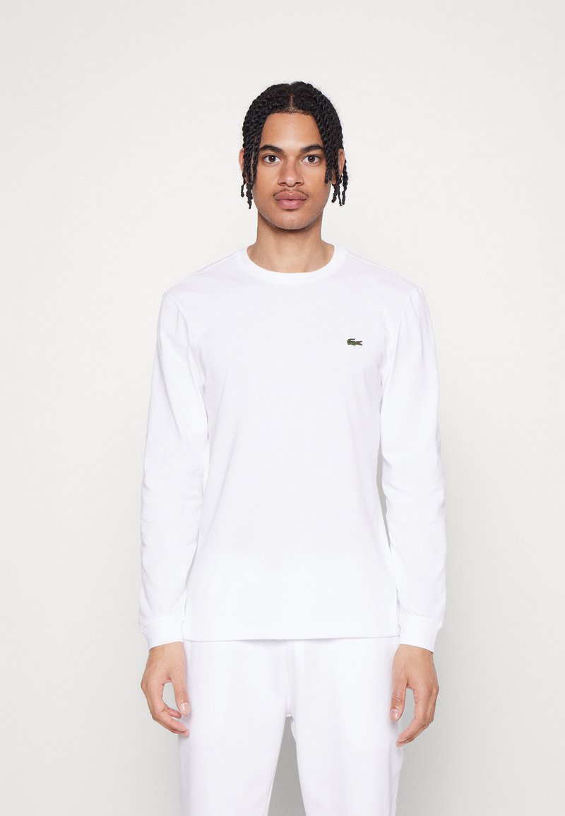 T-shirt blanc à manches longues en coton, avec un petit logo vert Lacoste sur la poitrine. Design simple à col rond. Texture douce.