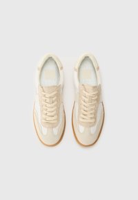 Des chaussures de sport beige avec un dessus en suede et cuir, des accents en forme de vagues, des lacets plats et une semelle en caoutchouc, dotées d'une semelle intérieure texturée.