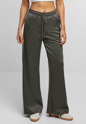 Femme portant un pantalon large en satin gris foncé avec des bandes blanches sur les côtés et des baskets blanches, debout devant un fond uni.