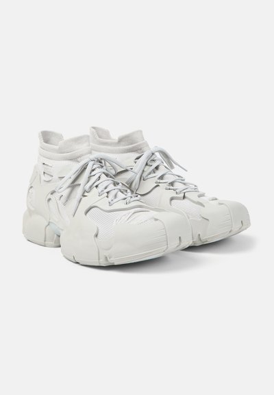Paire de baskets montantes blanches futuristes en tissu tricoté, semelles épaisses et lacets sur un fond clair uni.