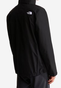 Personne portant une veste noire North Face vue de dos avec le logo de la marque sur la partie supérieure droite de l'épaule.
