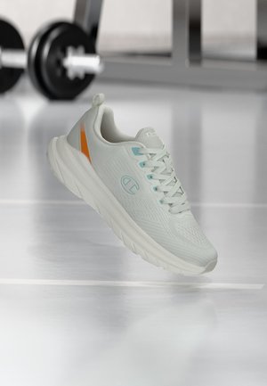 Scarpa da ginnastica grigio chiaro con accenti blu e arancioni che fluttua sopra un pavimento di palestra con manubri sfocati sullo sfondo.