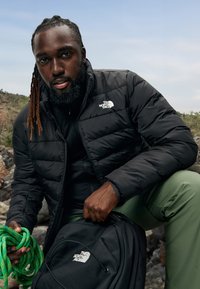 Chaqueta acolchada negra aislante con cierre de cremallera, combinada con una mochila negra. El modelo sostiene una cuerda de escalada verde y lleva puestos pantalones verdes.