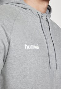 Sweat à capuche gris zippé en matériau doux, avec un logo blanc sur la poitrine et des cordons de réglage pour la capuche.