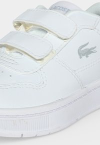 Άσπρο sneaker Lacoste με δύο λουριά βέλκρο και γκρι λογότυπο κροκόδειλου στο πλάι, εμφανίζοντας λεπτομερή ραφές και υφή σόλας.