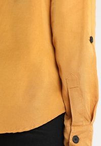 Långärmad skjorta i senapsgul tyg med slät textur. Har en figurnära manschett med en svart knapp och vinklad manschettdesign.