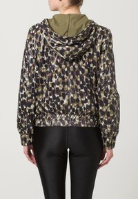 Hupparillinen takki, jossa on kiiltävä, kuvioitu kangas vihreänä, mustana ja ruskeana; vyötärö ja hihansuut kuminauhalla kiristettyjä; päällä mustat kiiltävät leggingsit.