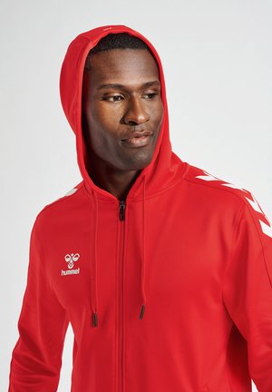 Rode zip-up hoodie van glad materiaal met een trekkoordkap. Heeft witte chevronpatronen op de schouders en een logo op de borst.