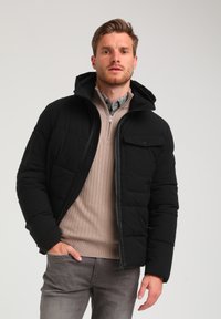 Sort pufferjakke med hætte, med en frontlynlås og en brystlomme. Sammen med en beige ribbet sweater og grå jeans.