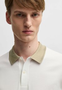 Polo bianco con colletto lavorato beige, dotato di una chiusura frontale a bottoni e una texture di cotone liscia. Ritratto di un modello maschile.